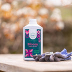 Bloom 1L bottle on a wooden table with a pair of purple gardening gloves and blossom behind
