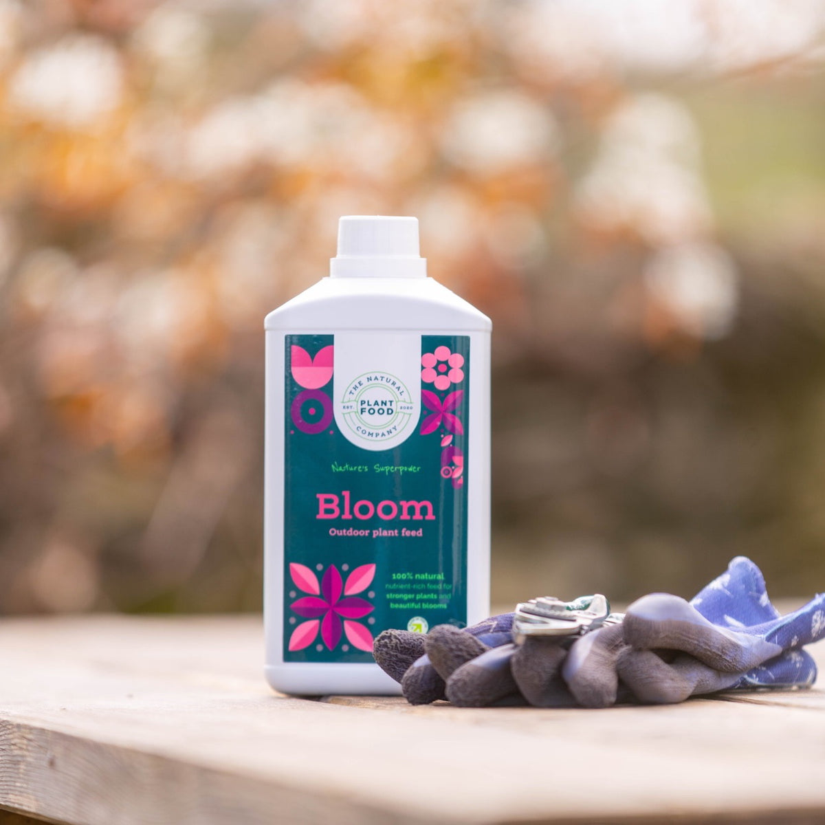Bloom 1L bottle on a wooden table with a pair of purple gardening gloves and blossom behind