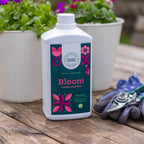 Bloom 1L bottle on a wooden table beside white pots of flowers, with gardening gloves and secateurs