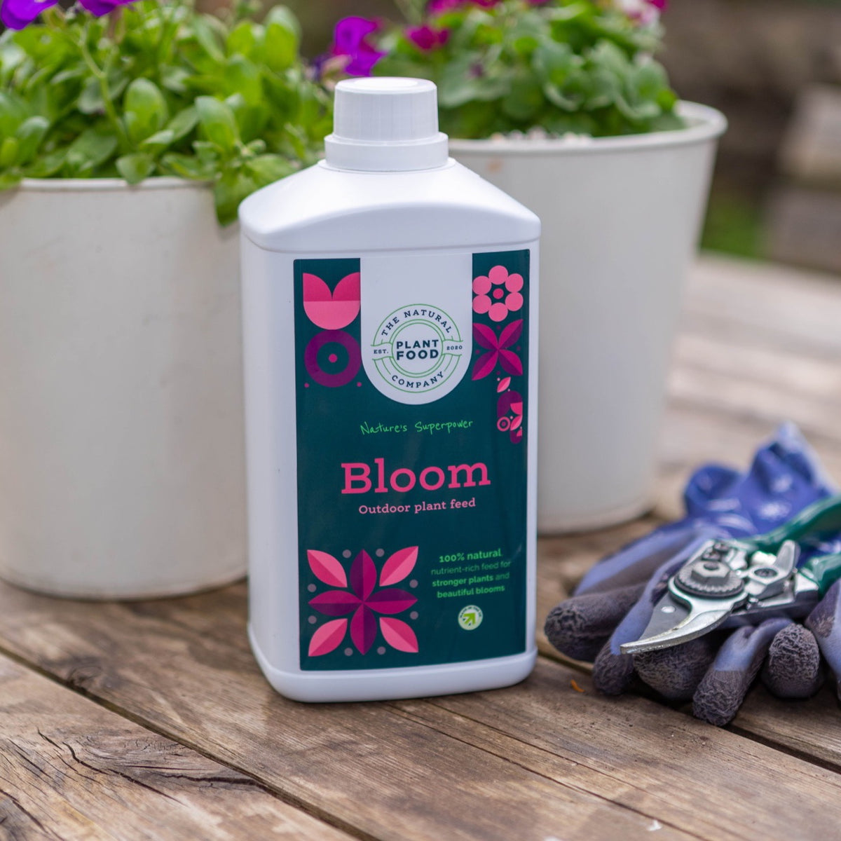 Bloom 1L bottle on a wooden table beside white pots of flowers, with gardening gloves and secateurs