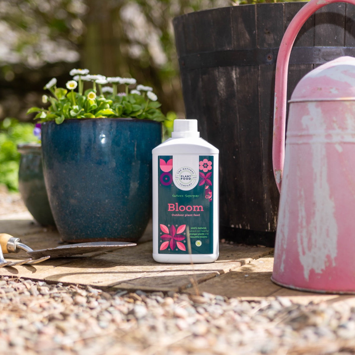 Bloom 1L bottle on a gravel path beside a blue glazed pot of daisies, a pink watering can and a hand trowel