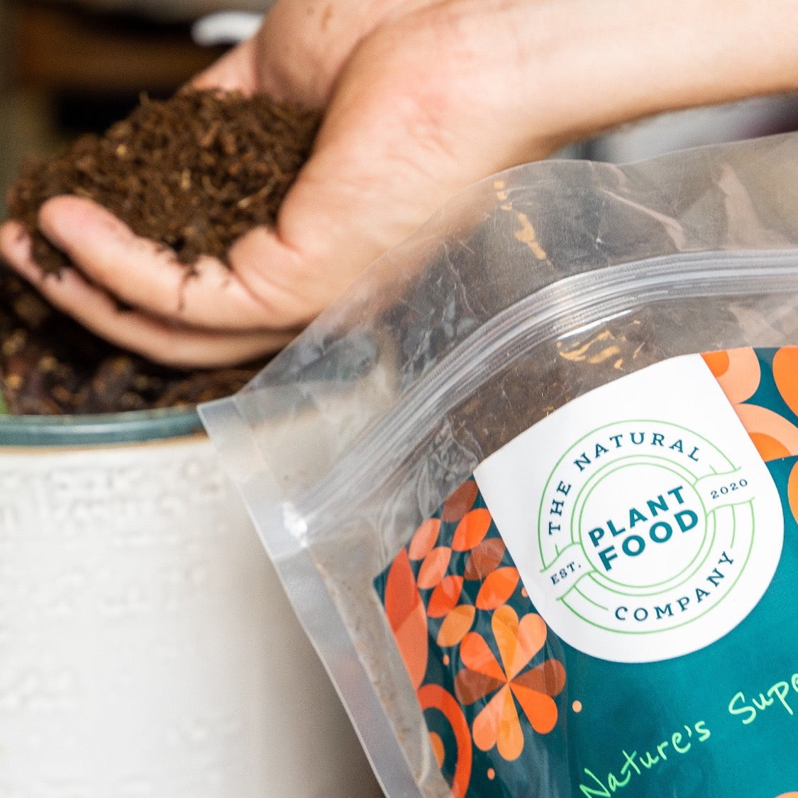 A hand holding a scoop of dark soil improver above the clear Soil Improver pouch