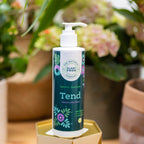 Close view of the Tend 250ml pump bottle on a gold plinth, with hydrangea flowers and plant pots out of focus behind